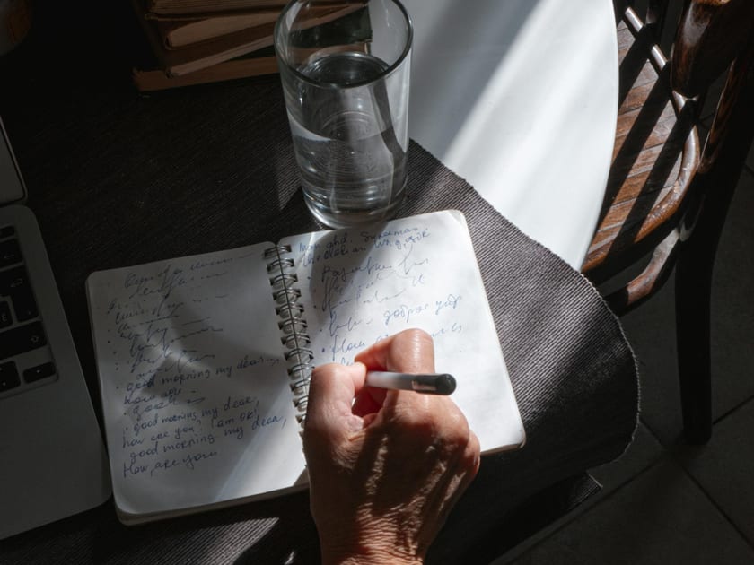 A person writing in a notebook with books and water nearby
