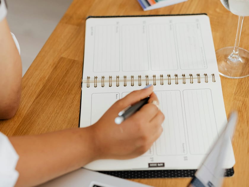 Person writing in a planner beside a laptop and glass of water